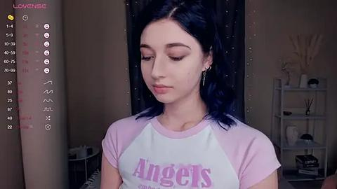 Media: Video of a young woman with fair skin and dark hair, wearing a pink and white T-shirt with \"Angels\" in purple. She has a serene expression. The background features a minimalist room with a white shelf and a curtain.