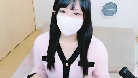 Media: Video of an Asian woman with long black hair, wearing a white face mask and pink cardigan, sitting on a beige couch in a minimalist room with a wall clock.