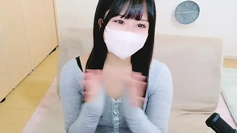 Media: A video of an East Asian woman with long black hair and pale skin, wearing a white mask and light blue long-sleeve top, sitting on a beige couch with a round clock on the wall behind her.