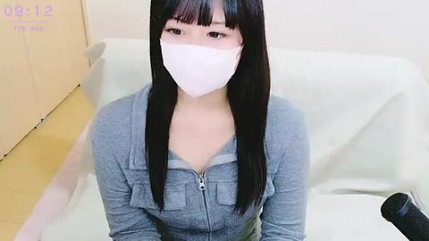 Media: Video of an East Asian woman with long black hair and pale skin, wearing a light gray sweater and white face mask, sitting on a white chair in a sterile medical room.