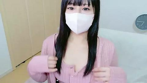 Media: Video of an East Asian woman with long black hair, wearing a pink cardigan and white mask, giving thumbs-up in a white-tiled bathroom with a clock on the wall.