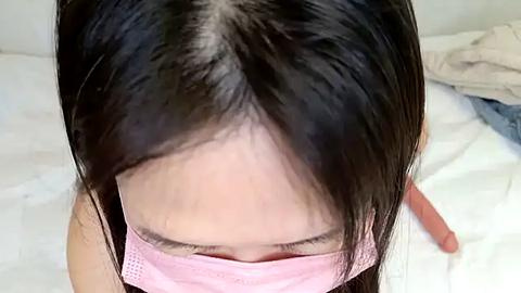 Media: Video of a young woman with straight black hair, wearing a pink face mask, looking downward. The background features a white bed and a partly visible blue shirt. The image is intimate and focused on her face.