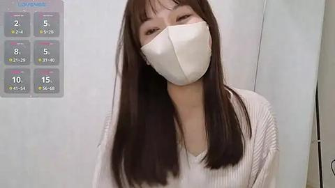 Media: Video of a young East Asian woman with straight, long black hair and fair skin, wearing a white ribbed sweater and white face mask. Background shows a white wall with a COVID-19 daily case chart.