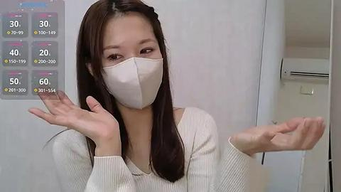 Media: Video of an Asian woman with straight, dark hair, wearing a white face mask and a light sweater, gesturing with both hands. Background features a white wall and a digital display showing temperature and humidity levels.