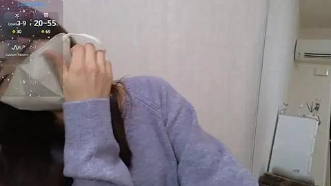 Media: Video of a person in a light purple sweater, wearing a clear plastic visor, holding a cup. Background features a white wall and an air conditioning unit.