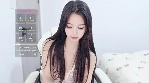 Media: Video of a young Asian woman with long, straight black hair, wearing a white tank top, sitting on a chair in a minimalist, white-walled room.