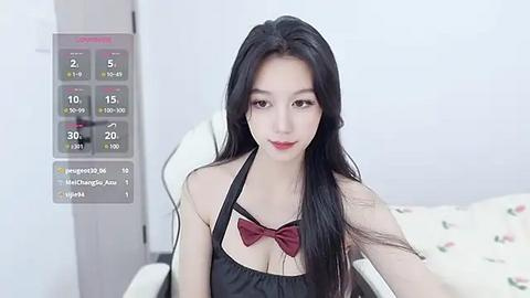 Media: Video of a young East Asian woman with long black hair, fair skin, and red lipstick, wearing a black apron with a red bow, seated on a bed with floral bedding.