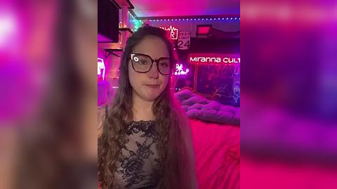 Media: Video of a smiling young woman with curly brown hair, wearing black-framed glasses and a patterned black top, standing in a dimly lit room with neon lights and a plush bed.