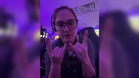 Media: A video shows a woman with glasses and short hair making a devil horn gesture inside a dimly lit room with purple lighting, blurred figures in the background.