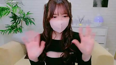 Media: Video of an Asian woman with long, wavy brown hair, wearing a black crop top and white face mask, making peace signs, in a modern, minimalist room with white walls, a potted plant, and a white dresser.