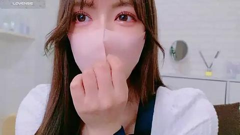 Media: Video of a young woman with fair skin and long brown hair, wearing a white towel over her head, covering her mouth and nose, looking towards the camera with large, expressive eyes.