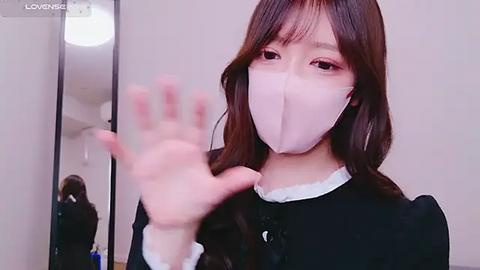 Media: A video of an Asian woman with long brown hair, wearing a face mask, black dress, and holding up a hand in front of a mirror.