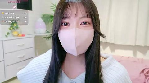 Media: Video of an East Asian woman with long black hair, wearing a white face mask, light gray sweater, in a cozy, softly lit bedroom with white furniture and green plants.