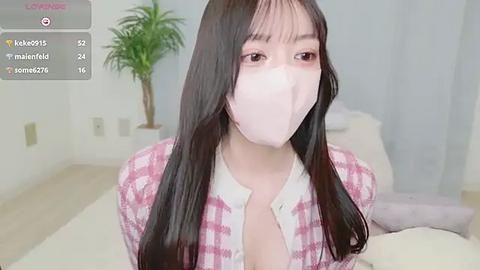 Media: Video of a young East Asian woman with long black hair, wearing a pink plaid shirt, white mask, and sitting indoors on a bed with white sheets.