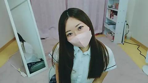 Media: Video of an Asian woman with long brown hair, wearing a light blue shirt and pink face mask, sitting in a room with a mirror, pink curtains, and cluttered shelves.