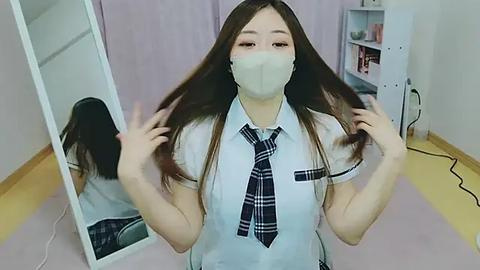 Media: A video of an East Asian woman with long, straight brown hair, wearing a white shirt, plaid tie, and a face mask, standing in a pastel-colored room with a mirror and shelves.