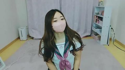 Media: Video of a young Asian woman with long brown hair, wearing a pink surgical mask, a sailor-style school uniform, and standing in a sparsely furnished room with white walls, a light purple curtain, and a white shelf.