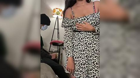 Media: A video shows a woman with long dark hair in a leopard-print dress, standing in a dimly lit room with a lamp and a chair in the background.