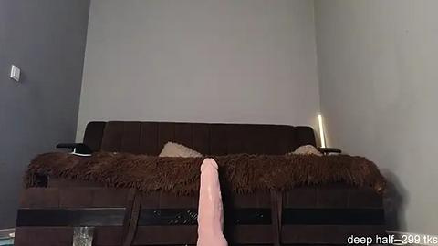 Media: Video of a dark brown, tufted sofa with a fluffy brown throw, a pink towel hanging off the edge, and a lit candle on the right side in a minimalist, beige-walled room.