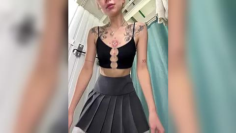 Media: A video of a tattooed woman in a black crop top and pleated gray skirt, standing in a bathroom with a teal shower curtain and white wall.