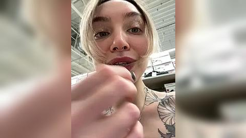 Media: A close-up video of a blonde woman with a nose ring and tattoos, wearing a black bra, in a warehouse setting.