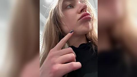 Media: Video of a young woman with long blonde hair, wearing a black top, looking up thoughtfully, with her fingers touching her nose, and a blurred figure in the foreground.