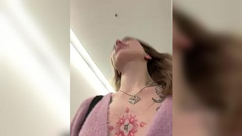 Media: Video of a woman with fair skin and long brown hair, wearing a pink cardigan and silver necklace, displaying a detailed red and black tattoo on her chest.