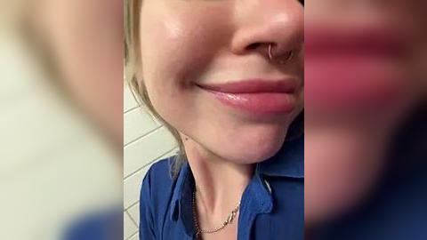 Media: Video of a close-up of a smiling woman with fair skin, wearing a blue shirt, nose ring, and shiny pink lipstick.