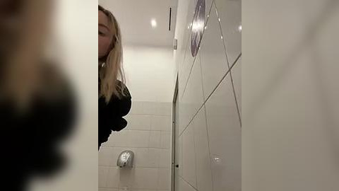 Media: A video of a blonde woman in a bathroom, wearing a black top, standing in front of a white-tiled shower with a silver faucet and handle.