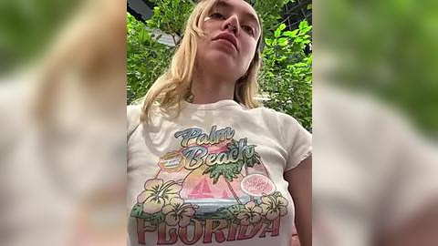 Media: Video of a young, fair-skinned woman with long blonde hair, wearing a colorful \"Paradise Beach\" T-shirt, standing against lush green foliage.