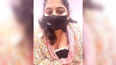 Media: Video of a South Asian woman with medium brown skin, wearing a black face mask, floral blouse, and pink shawl, sitting against a blurred, light-colored background.