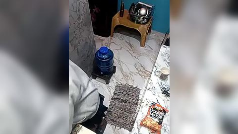 Media: A video showing a person in a white shirt and black pants standing near a blue water cooler, a small wooden stool, and a stack of newspapers on a tiled floor.