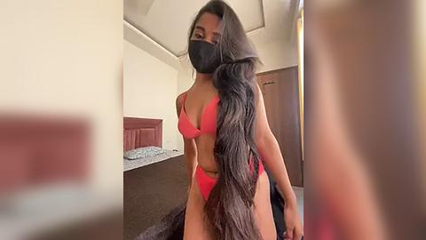 Media: Video of a slender, light-skinned woman with long, black hair wearing a red bra and panties, a black face mask, and a black jacket, standing in a dimly lit bedroom with a bed, wooden door, and beige walls.