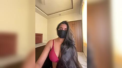 Media: Video of a woman with long black hair, wearing a pink bra and black face mask, seated in a dimly lit, beige-walled room with a wooden door and a marble countertop.