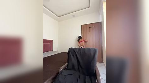 Media: A video of a woman in a red bra, sitting on a bed with a black blanket, in a minimalist, beige-walled room with a wooden door and a small window.