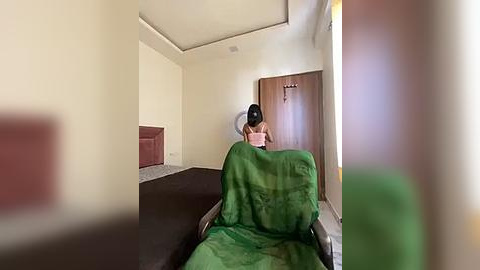 Media: A video of a sparse, beige-walled bedroom with a green chair, a wooden door, and a bed with a dark brown blanket.