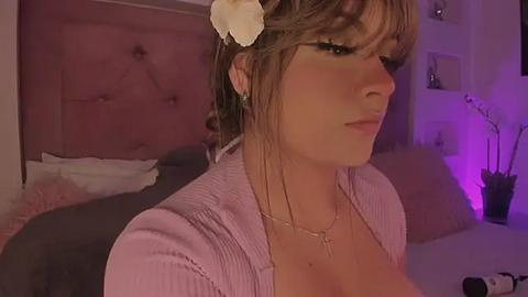 Media: Video of a fair-skinned woman with light brown hair, wearing a pink ribbed cardigan, a dainty necklace, and a white flower headpiece, standing in a dimly-lit bedroom with pink walls, a bed, and a purple light.