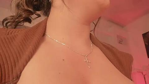 Media: Video of a woman with fair skin and dark hair, wearing a brown ribbed sweater, and a delicate silver necklace with a small pendant. Background shows a dimly lit, pink-lit room with a ceiling fan.