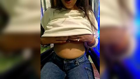 Media: Video of a young woman lifting her white T-shirt to expose her midriff and blue jeans, sitting in a dimly lit room with a blurred background.