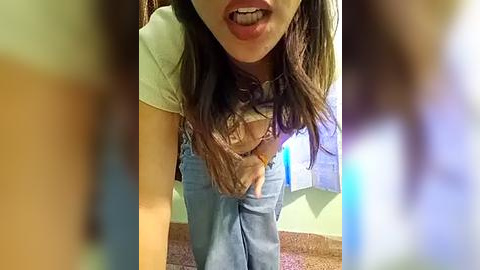 Media: Video of a young woman with long brown hair, wearing a white t-shirt and blue overalls, sticking out her tongue, standing in a carpeted room with pastel walls and a blue poster.