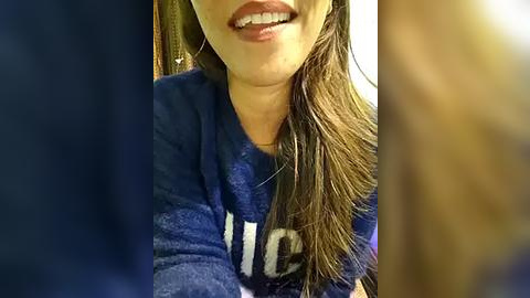 Media: A close-up video of a woman with medium skin tone and straight, shoulder-length brown hair, smiling with braces. She wears a blue sweatshirt with a partially visible white graphic. The blurred background includes a wooden door and a fuzzy, light-colored object.