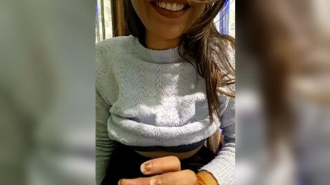 Media: Video of a smiling woman with long brown hair, wearing a light blue, textured knit sweater, and a black skirt. Her hands are visible, with a watch on her wrist. Background shows blurred indoor setting with white walls.