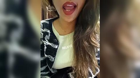 Media: Video of a young woman with long brown hair, wearing a white t-shirt and a black-and-white patterned blazer, sticking out her tongue playfully.