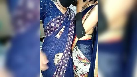 Media: A video of two women in blue sarees with intricate patterns, wearing black masks, standing close together indoors. The background is blurred, focusing on the sarees and their close proximity.