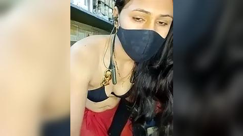 Media: Video of a woman with long black hair, wearing a black face mask, red dress, and gold necklace, leaning forward, taken from a low angle.