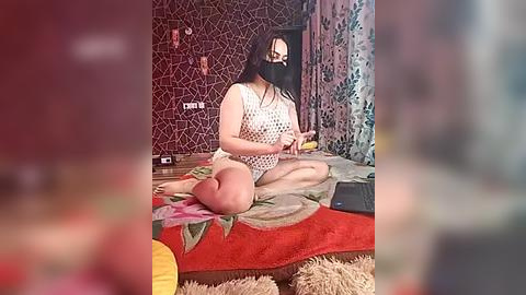 Media: Video of a fair-skinned woman with long dark hair, wearing a white sleeveless polka-dot dress, black mask, and sitting cross-legged on a bed, surrounded by red and green bedding, in a dimly lit room with intricate wallpaper.