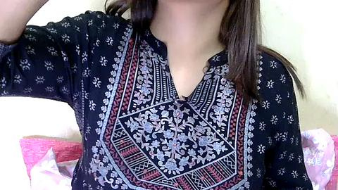 Media: Video of a woman with medium skin tone, wearing a black long-sleeve shirt adorned with intricate, geometric, and floral embroidery in shades of gray, white, and red. Her long, dark brown hair cascades down her back.