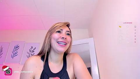 Media: A video of a smiling, light-skinned woman with straight, shoulder-length blonde hair, wearing a black tank top. She's in a room with a white ceiling, a calendar, and a closed door in the background.