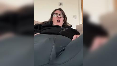 Media: A video captures a woman in a black hoodie, sitting with legs spread, wearing black leggings, and glasses, appearing surprised.