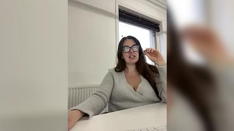 Media: A video of a curvy woman with long brown hair, glasses, and a gray sweater, adjusting her glasses while sitting at a white table in a bright, minimalistic room with a white radiator and window.